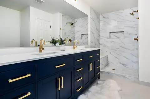 Primary bathroom with a navy double vanity, brass fixtures and a marble-look tiled walk-in shower
